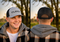 Front-and-back portrait of a smiling man in a gray hoodie and plaid overshirt wearing the gray and black FlekX Classic Hat at dusk; the structured gray front carries bold black FLEKX Performance Fishing embroidery, the rear view black mesh with an adjustable snap closure. A christian fishing trucker hat from a veteran owned fishing apparel brand.