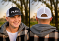 Smiling young man in a gray hoodie and plaid overshirt wearing the black-front FlekX Classic Hat at dusk; the front view shows crisp white FLEKX Performance Fishing embroidery on the structured crown, the rear view white breathable mesh with an adjustable snap closure. A christian fishing trucker hat from a veteran owned fishing apparel brand.